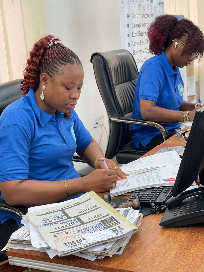 Two women in casual office attire working at desks in a business setting, reviewing documents.